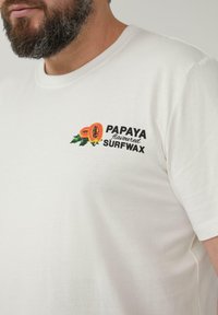 Witte katoenen t-shirt met een graphic van papaja's en de tekst "PAPAYA smaak SURFWAX." Korte mouwen en ronde hals. Zachte textuur.