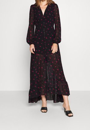 Kvinde iført en sort, langærmet maxi-kjole med knapper og røde blomsterprint samt sorte hælede slippers mod en hvid baggrund.