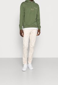 Muž v olivově zelené mikině s kapucí, bílém tričku, béžových kalhotách a bílých teniskách stojí před jednoduchým světle šedým pozadím.