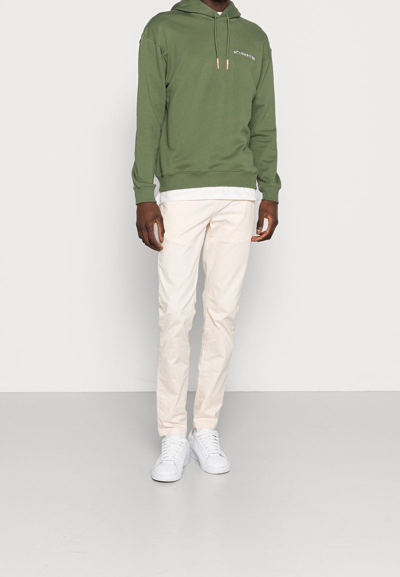 Muž v olivově zelené mikině s kapucí, bílém tričku, béžových kalhotách a bílých teniskách stojí před jednoduchým světle šedým pozadím.