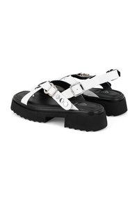 Witte platte sandalen met een dikke zwarte zool, voorzien van gespsluiting en metalen accenten. Gekruiste banden zorgen voor een goede pasvorm.