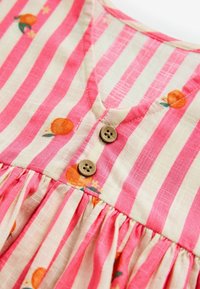 Tissu rayé rose et blanc avec des imprimés d'oranges, deux petits boutons marron et des fronces sous la poitrine.
