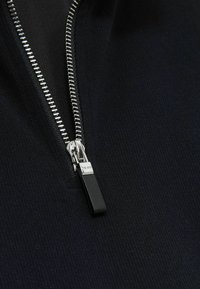 Prenda de color negro con cremallera que presenta una tela texturizada. Cremallera de metal plateado con un tirador de cuero negro y una cinta de cremallera suave.