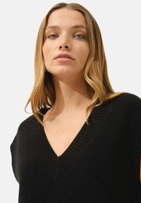 Svart v-ringad ärmlös tröja med ribbat mönster och axeldetaljer, tillverkad av mjukt tyg och med en avslappnad passform.