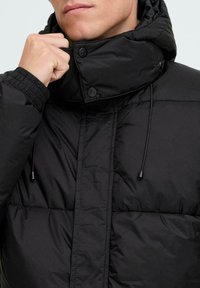 Melns pūkains jaka ar augstu apkakli, spiedpogām, regulējamu auklu un elastīgām piedurknēm. Tekstūrēta auduma ar quilted rakstu.