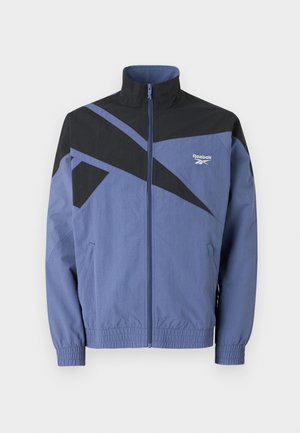 Reebok windbreaker in blue and black, featuring a high collar, full zip, elastic cuffs, and angled color-block design with logo.