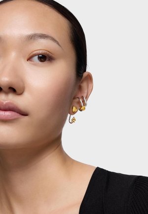 Zbliżenie na lewe ucho kobiety, na którym znajdują się trzy srebrno-złote kolczyki: dwa w kształcie serca oraz jeden w kształcie strzały.