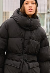 Svart puffjacka med hög krage, framknäppning och midjerem. Har strukturerad tygkvalitet och sidofickor.