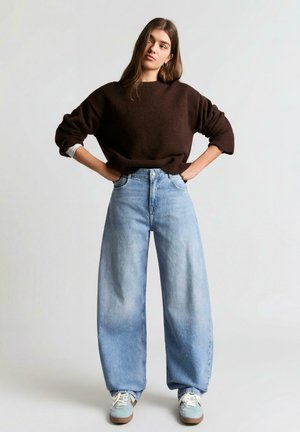 Tânără stă cu mâinile pe șolduri, purtând un pulover maro închis, blugi largi de culoare albastru deschis și adidași albi, pe un fundal simplu.