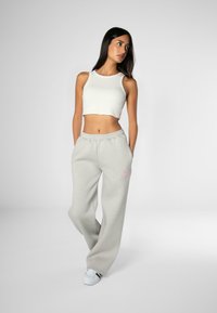 White cropped tank top and light grey sweatpants with elastic waistband and pink logo. Model stands casually against a neutral background.
