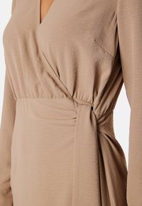Beżowa sukienka kopertowa z miękkiego, lekkiego materiału, z głębokim dekoltem w kształcie litery V i węzłem z boku, z długimi rękawami oraz zwiewnym krojem.