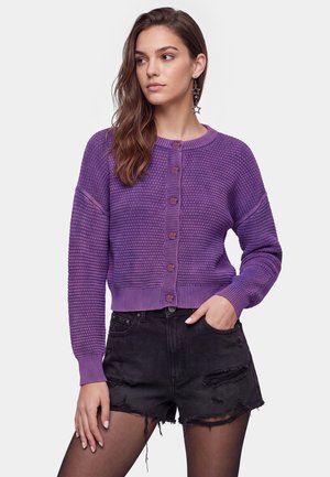 Kvinde iført en lilla strikket cardigan med knapper, sorte slidte denimshorts, sorte strømpebukser og stjerneformede hængende øreringe, stående med den ene hånd i lommen.