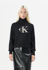 Zwarte gebreide trui met een hoge nek en ribbels aan de mouwen. Kenmerkt zich door een groot "CK"-logo in lichtgrijs op de voorkant, gecombineerd met een zwarte faux leren rok.