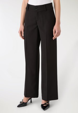 Pantaloni neri a gamba larga con una texture liscia, caratterizzati da due bottoni in vita. Abbinati a décolleté neri in vernice.