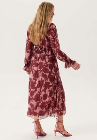 Kvinna med långt blont hår som bär en långärmad, rosa och vinröd blommönstrad midiklänning och rosa klacksandaler, står med ryggen vänd.