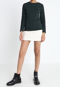 Haut à manches longues vert foncé avec des manches texturées, associé à une mini-jupe blanche. Le mannequin porte des bottines noires avec des panneaux élastiques sur les côtés.