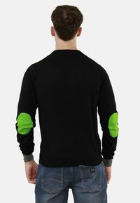 Maglietta nera a maniche lunghe con toppe verdi sui gomiti, polsini grigi e design aderente, vista da dietro.