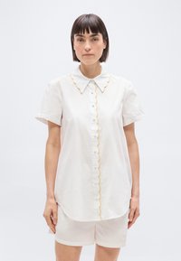 Femme aux cheveux longs et raides, portant un chemisier blanc à manches courtes avec un bord festonné et un short blanc assorti, debout devant un fond uni.