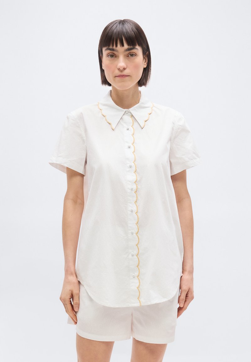 Femme aux cheveux longs et raides, portant un chemisier blanc à manches courtes avec un bord festonné et un short blanc assorti, debout devant un fond uni.