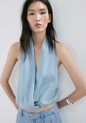 Jeune femme aux longs cheveux foncés portant un haut drapé bleu clair sans manches et un pantalon en jean assorti, debout contre un mur blanc uni.