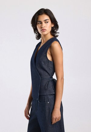 Vrouw met kort donker haar die een mouwloos marineblauw topje met krijtstreep en bijpassende broek draagt, kijkt naar links tegen een effen achtergrond.
