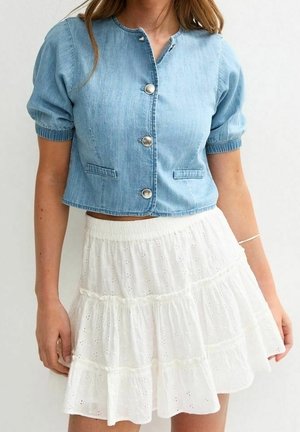 Femme portant une veste en denim bleu clair à manches courtes et boutonnée, et une jupe blanche à volants en broderie anglaise.