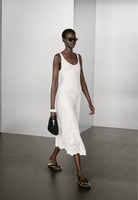 Witte mouwloze bodysuit gecombineerd met een kanten rok met ingewikkelde patronen. Accessoires zijn onder andere donkere zonnebril en een zwarte gestructureerde tas.