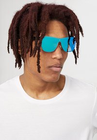 Zilas aplokšņu saulesbrilles ar plakanu dizainu un atstarojošām lēcām, apvienotas ar baltu T-kreklu, ko valkā persona ar stilizētām matēm.