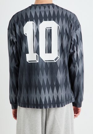 Personne portant une chemise à manches longues noire et grise à motif losange avec un grand numéro 10 blanc au dos, associée à un pantalon gris clair.