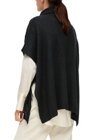Zwarte gebreide poncho met een hoge kraag, met een oversized vorm en zijslitsen, gedragen over een witte overhemd.