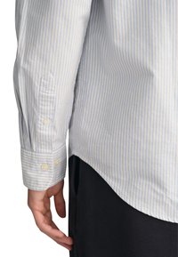 Camicia a righe in cotone azzurro chiaro e bianco. Presenta un orlo arrotondato, polsini con bottone e una fessura laterale per facilitare i movimenti.