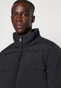 Veste épaisse noire en quilté avec un col montant, équipée d'une fermeture éclair à l'avant et d'un petit logo sur le côté gauche. Texture lisse.