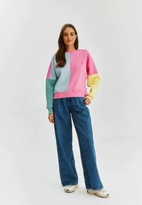 Kolorowy sweatshirt w odcieniach różu, niebieskiego, zielonego i żółtego, o luźnym kroju i ściągaczami. Stylizowany z szerokimi niebieskimi dżinsami i trampkami.