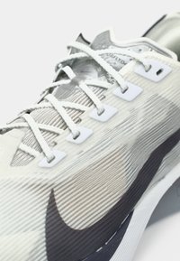 Prim-plan al unei pantofi sport albi și gri, cu țesătură texturată, șireturi albe și un logo swoosh întunecat și proeminent pe partea laterală.