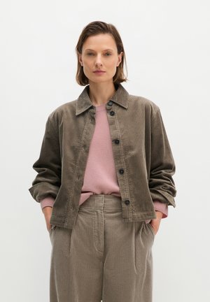 Vrouw draagt een bruine corduroy jas en broek met een roze trui, staand met handen in de zakken tegen een effen witte achtergrond.