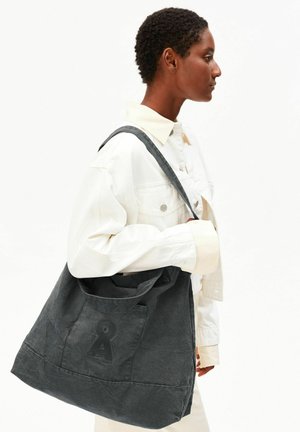 Personne en veste blanche porte un grand sac fourre-tout gris foncé avec un symbole de trou de serrure sur fond blanc uni.