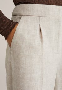 Käsi taskus heledates beežides kõrge vöökohaga pükstes, millel on voldid, koos pikkade varrukatega tekstuurse pruuni ülevalt.