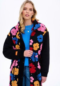 Čierna flísová bunda s farebnými kvetinovými vzormi v ružovej, modrej a oranžovej; má zips vpredu, cvoky a bočné vrecká.