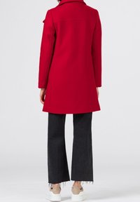 Cappotto rosso in lana con colletto strutturato e dettagli nella cucitura posteriore, abbinato a jeans neri a palazzo e sneakers bianche con accenti beige.