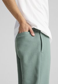 Pantalones deportivos verde claro con bolsillos laterales, hechos de una tela suave. Llevados con una camiseta blanca, que presenta un corte relajado y una textura suave.