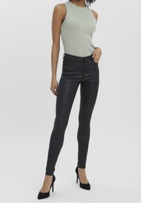 Jean noir slim associé à un haut sans manches côtelé vert clair. Le mannequin se tient en talons noirs classiques devant un arrière-plan neutre.