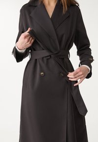 Schwarzer Trenchcoat aus glattem Stoff, mit doppelter Knopfleiste, Taillengürtel und großen Revers. Auffällige lange Ärmel mit geknöpften Manschetten.