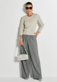 Frau trägt einen beigen Pullover mit gewelltem Saum, graue weit geschnittene Hose, große Sonnenbrille, silberne Creolen, Armbänder und hält eine silberne Handtasche.
