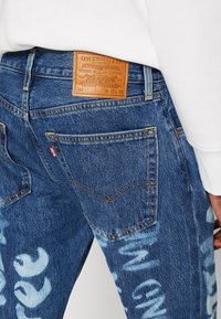 Pantalones vaqueros de mezclilla azul con una etiqueta de cuero en la cinturilla, que presentan costuras distintivas, bolsillos de solapa y patrones abstractos en azul claro en las piernas.