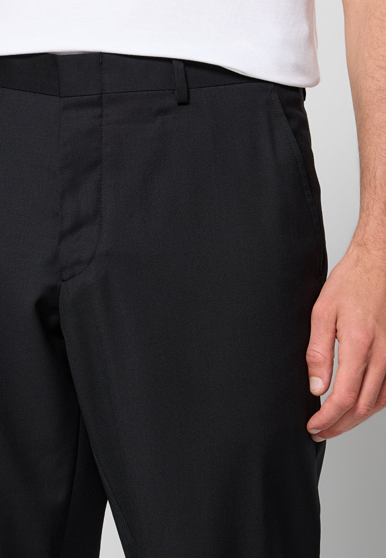 Man in zwart op maat gemaakte broek en een wit shirt, met de rechterhand ontspannen aan de zijkant, tegen een eenvoudige lichte achtergrond.