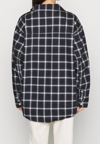 Person klädd i en lös svart- och vitrutig långärmad skjorta, sedd bakifrån, i kombination med ljusa byxor.