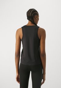 Widok z tyłu stojącej osoby z warkoczami, noszącej bezrękawnik w kolorze czarnym i czarne legginsy na jednolitym jasnym tle.