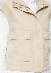 Gilet sans manches beige en tissu résistant, doté de deux poches avant, d'une fermeture éclair et de boutons-pression au col.