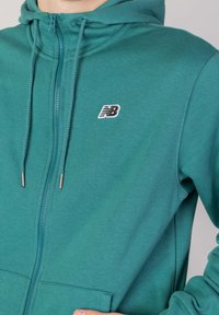 Turkos zip-up hoodie i mjukt tyg, med en framficka, dragsnoddshuva och broderad logga på bröstet.