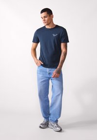 T-shirt à manches courtes bleu marine avec un petit logo blanc sur la poitrine, assorti à un jean en denim bleu clair et des chaussures de sport grises.